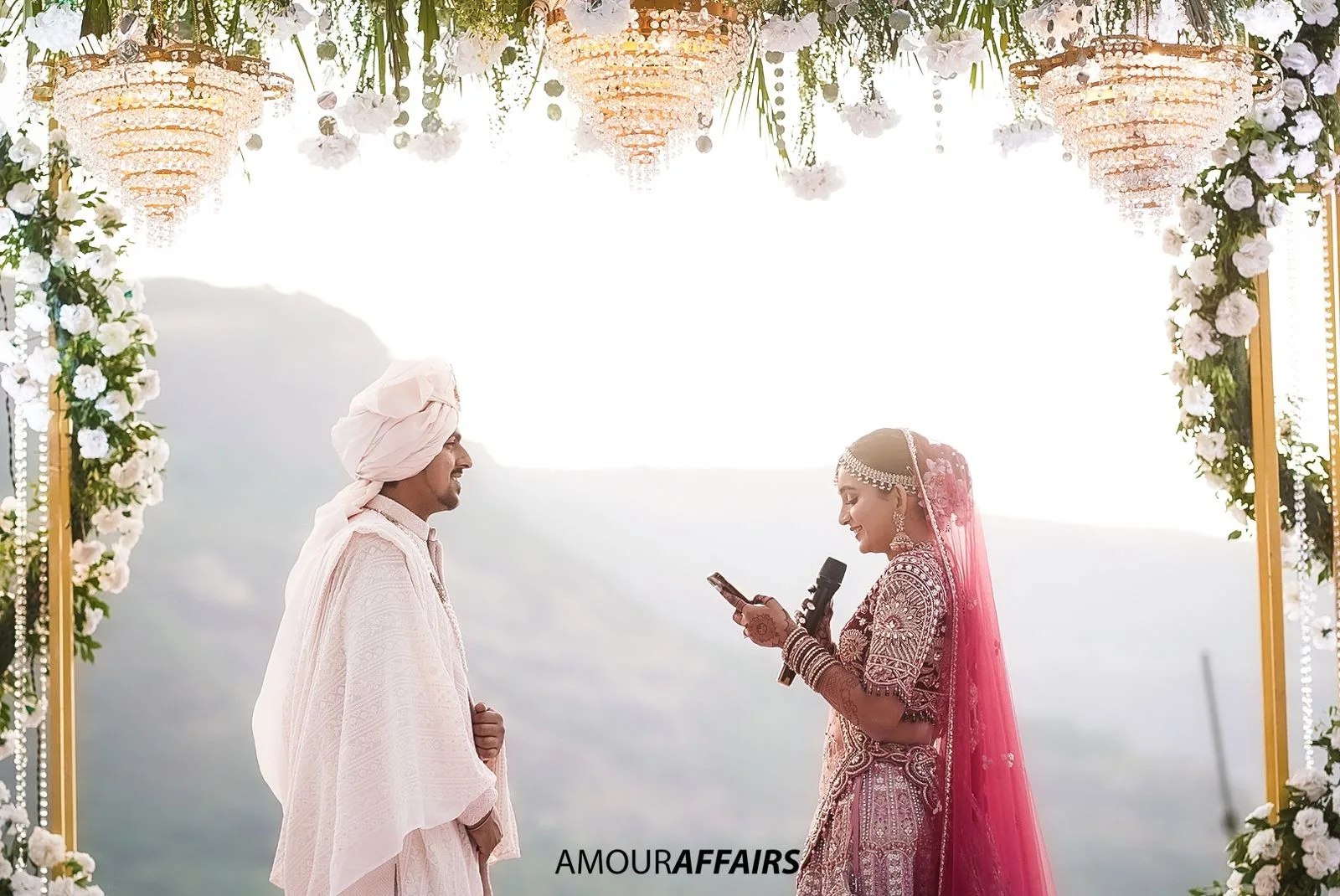 Candid wedding photograph of a bride reading her vows into a microphone while the groom listens at the mandap