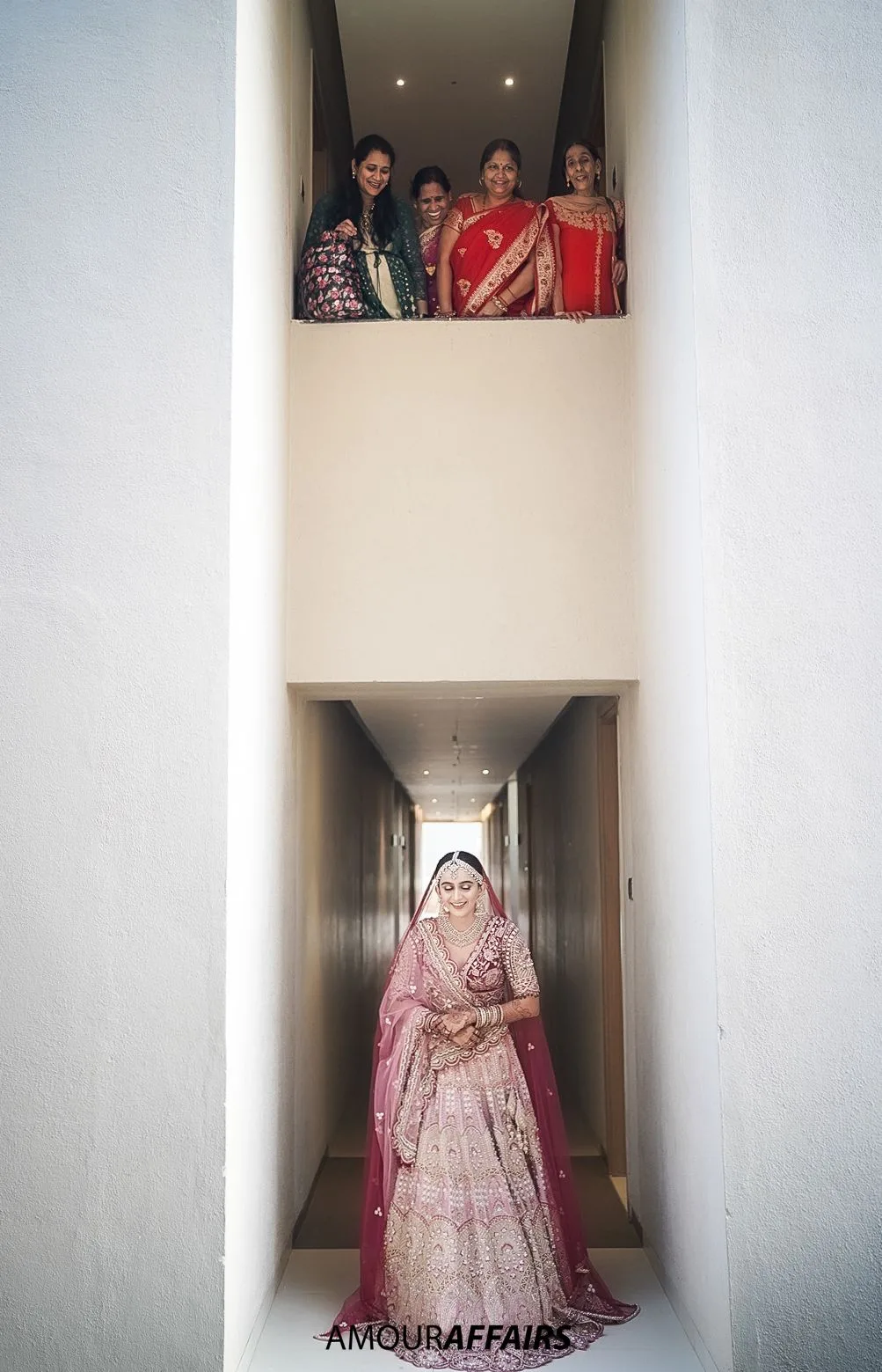 Bride in a pink lehenga walking down a corridor while the women of her family watch from a window above