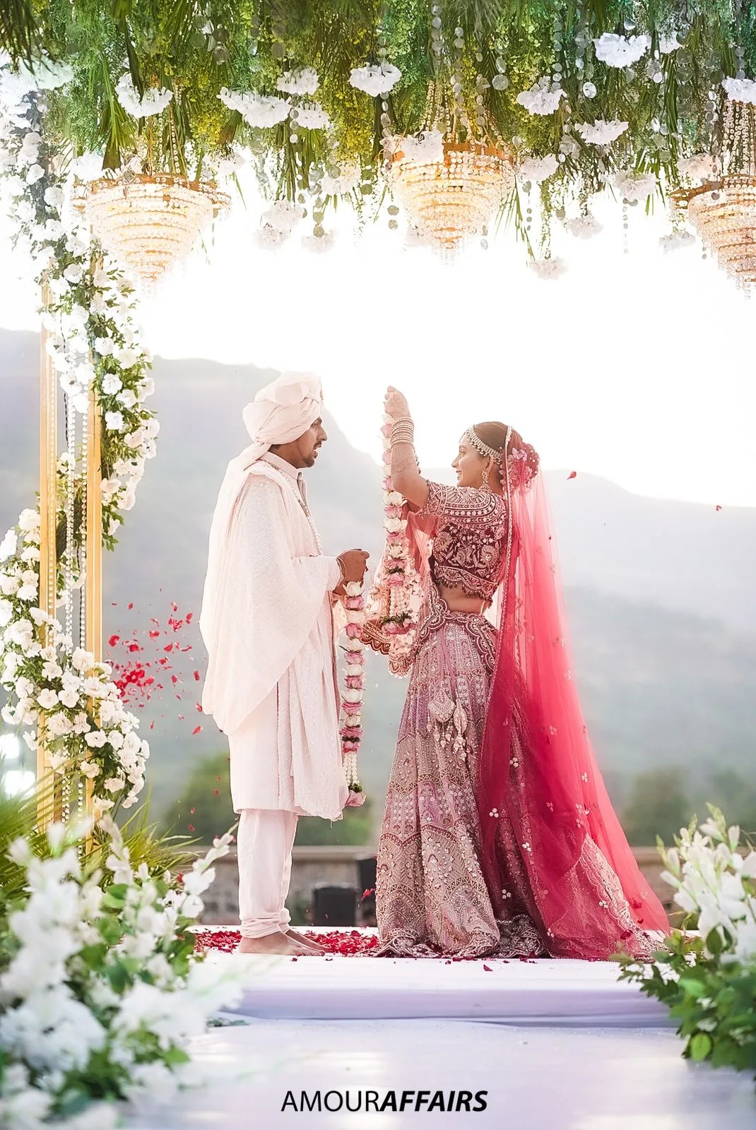 Bride and groom exchanging varmala garlands under a floral mandap as rose petals fall around them